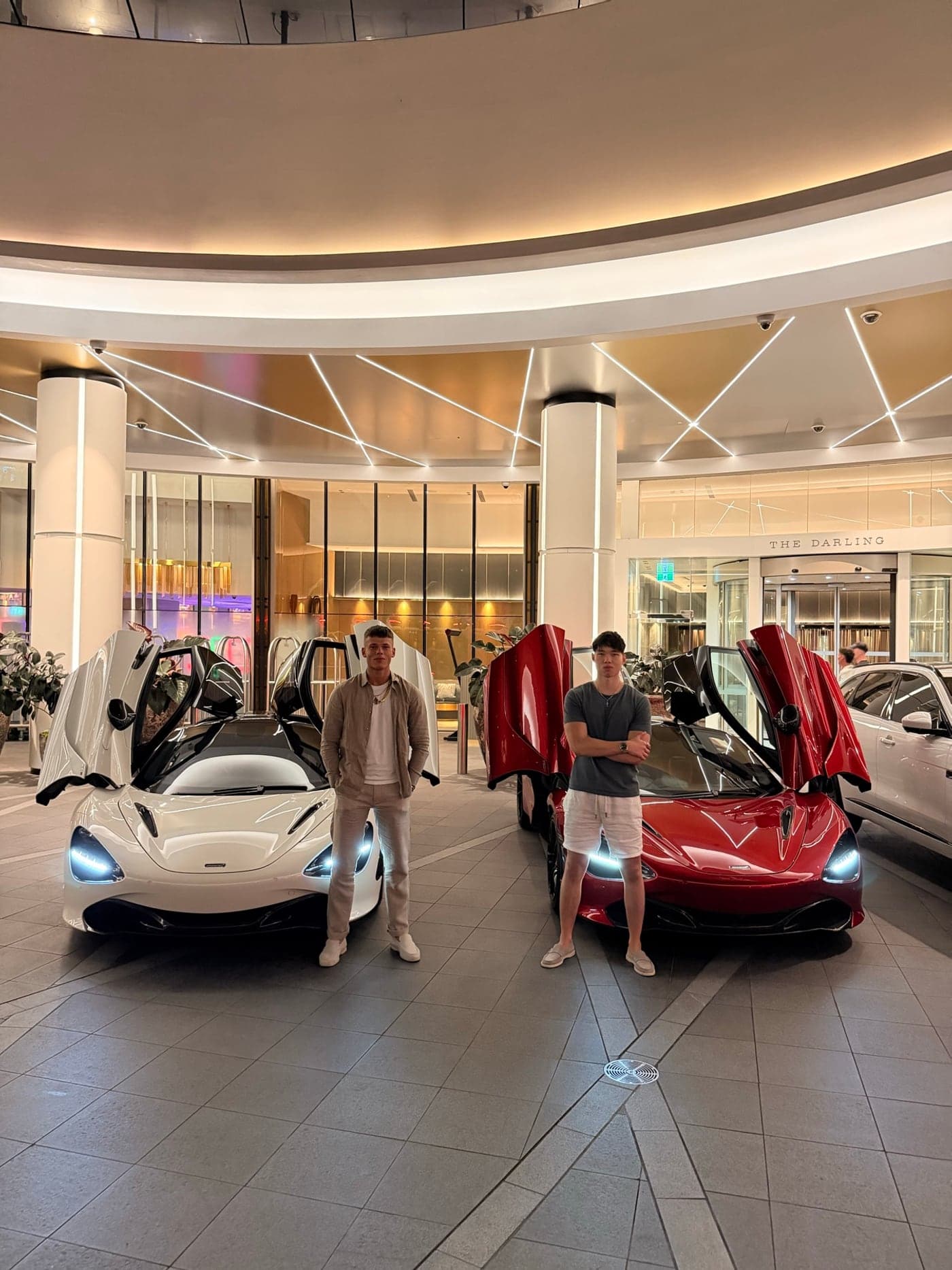 Max and Will standing in front of a white and a red McLaren 720S, doors up, in a hotel forecourt at night.